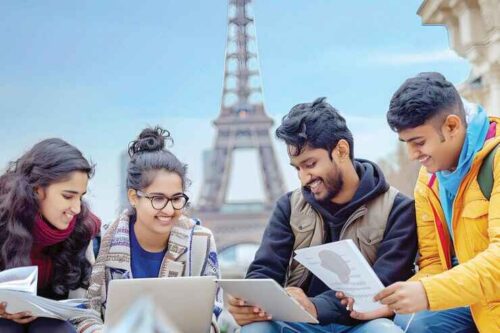 Four international students studying together near the Eiffel Tower in Paris, smiling and holding books and tablets.
